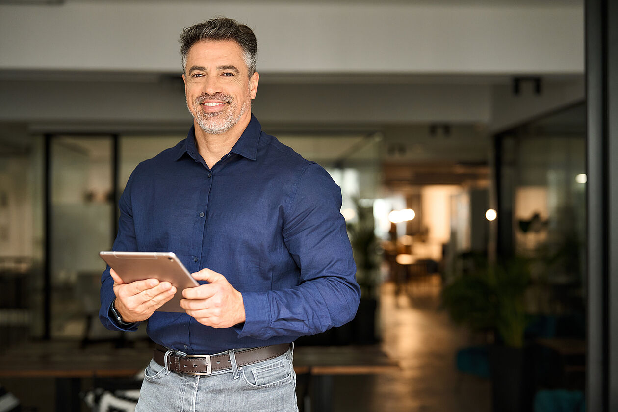 lächelnder Mann steht in einem modernen Büro und hält ein Tablet in den Händen